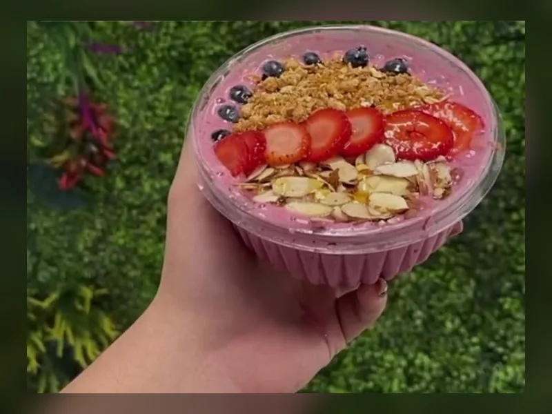 An açaí bowl topped with granola, strawberries, blueberries and sliced almonds, held up in front of the green wall at Fuel Up Energy Cafe in Bolingbrook, IL