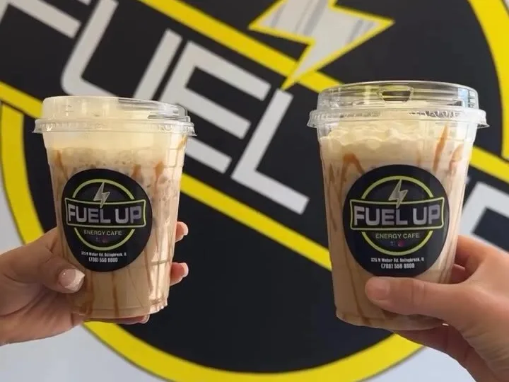 Two iced protein coffees with caramel drizzle raised in front of the Fuel Up logo wall at Fuel Up Energy Cafe in Bolingbrook